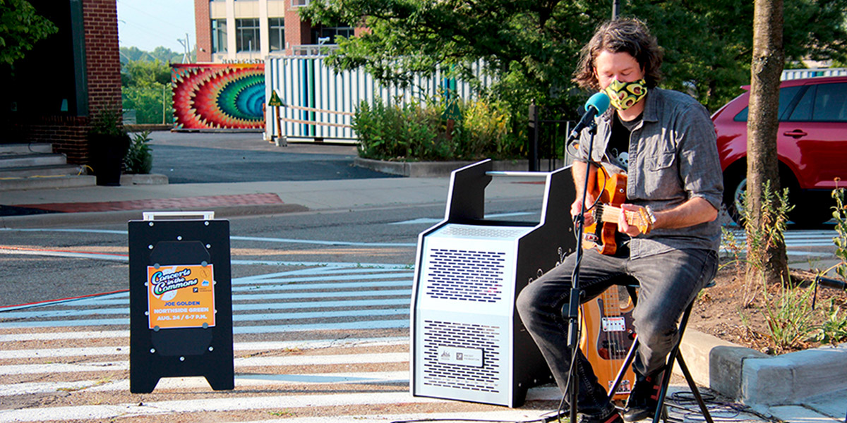 A man wearing a mask singing and playing guitar outside at the Earthquaker Devices Busking Station in the Northside District of downtown Akron.