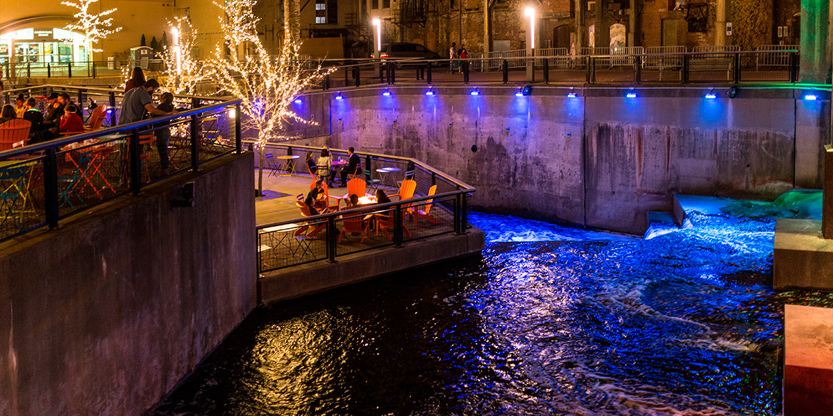 An image of Lock 4 in downtown Akron at night with colorful lights illuminating the water and white string lights in tree branches.