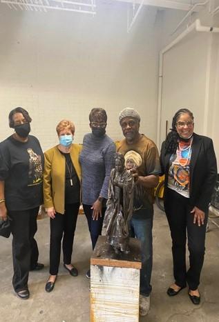 Four women and one man wearing surgical masks pose with a small prototype statue of Sojourner Truth in a room with a concrete floor and white walls